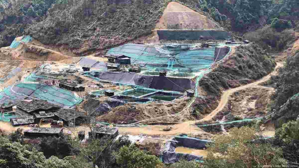 A view of a rare earth mining site surrounded by hills and forested terrain, showing excavation areas and processing pools.