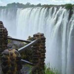 View of Victoria Falls from a wet stone walkway, showing water cascading over the cliff edge amid rising mist.