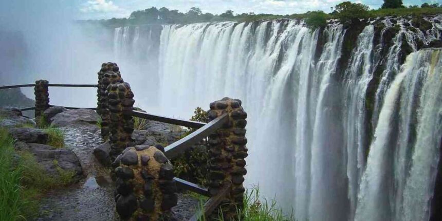 View of Victoria Falls from a wet stone walkway, showing water cascading over the cliff edge amid rising mist.