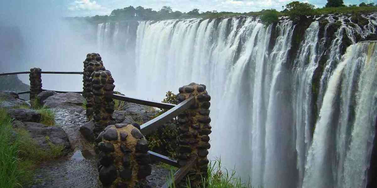 View of Victoria Falls from a wet stone walkway, showing water cascading over the cliff edge amid rising mist.